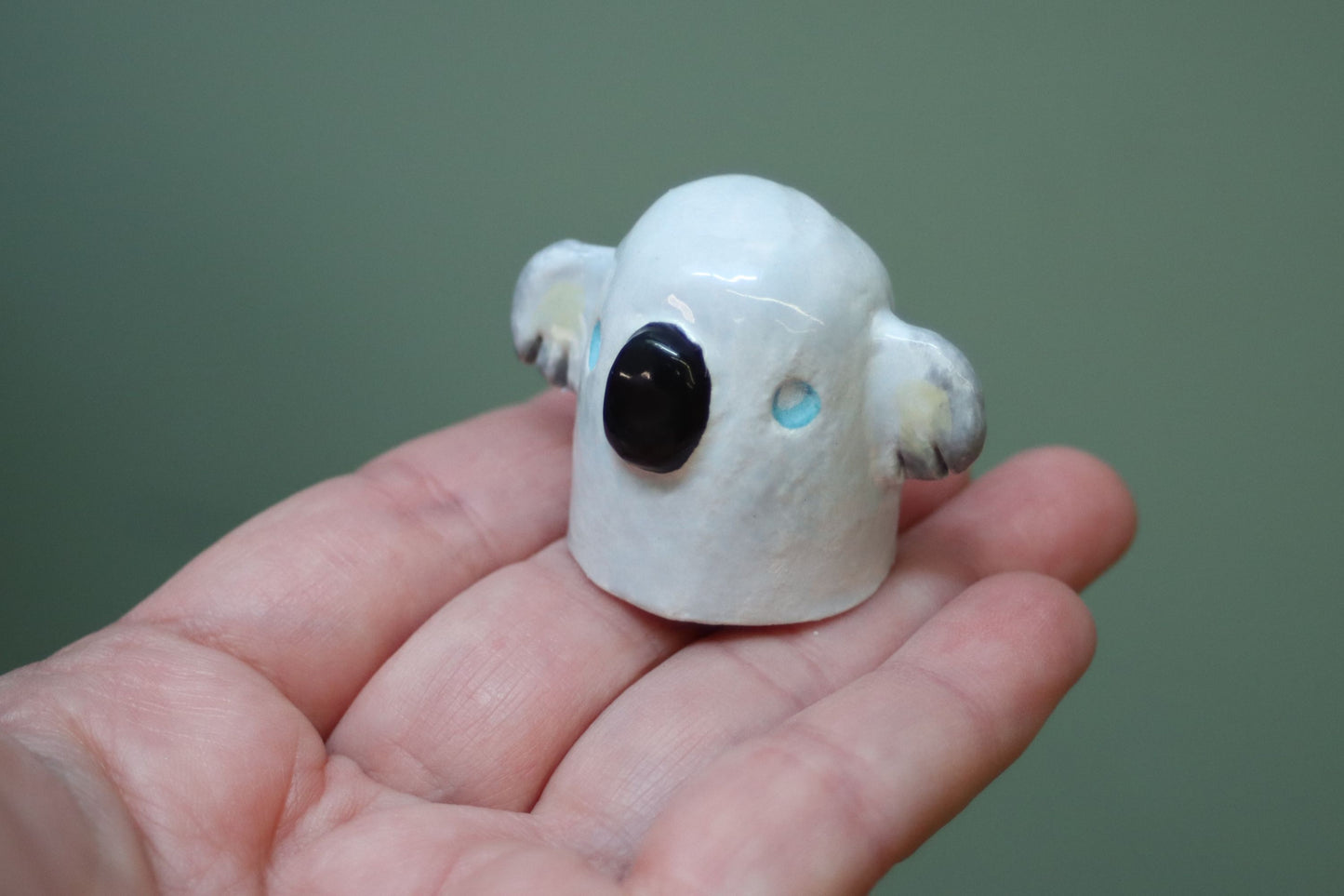 A small white koala figurine with a glossy finish, featuring a black nose, gray ears, and light blue eyes, held in a person's hand against a solid green background.