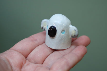 A small white koala figurine with a glossy finish, featuring a black nose, gray ears, and light blue eyes, held in a person's hand against a solid green background.